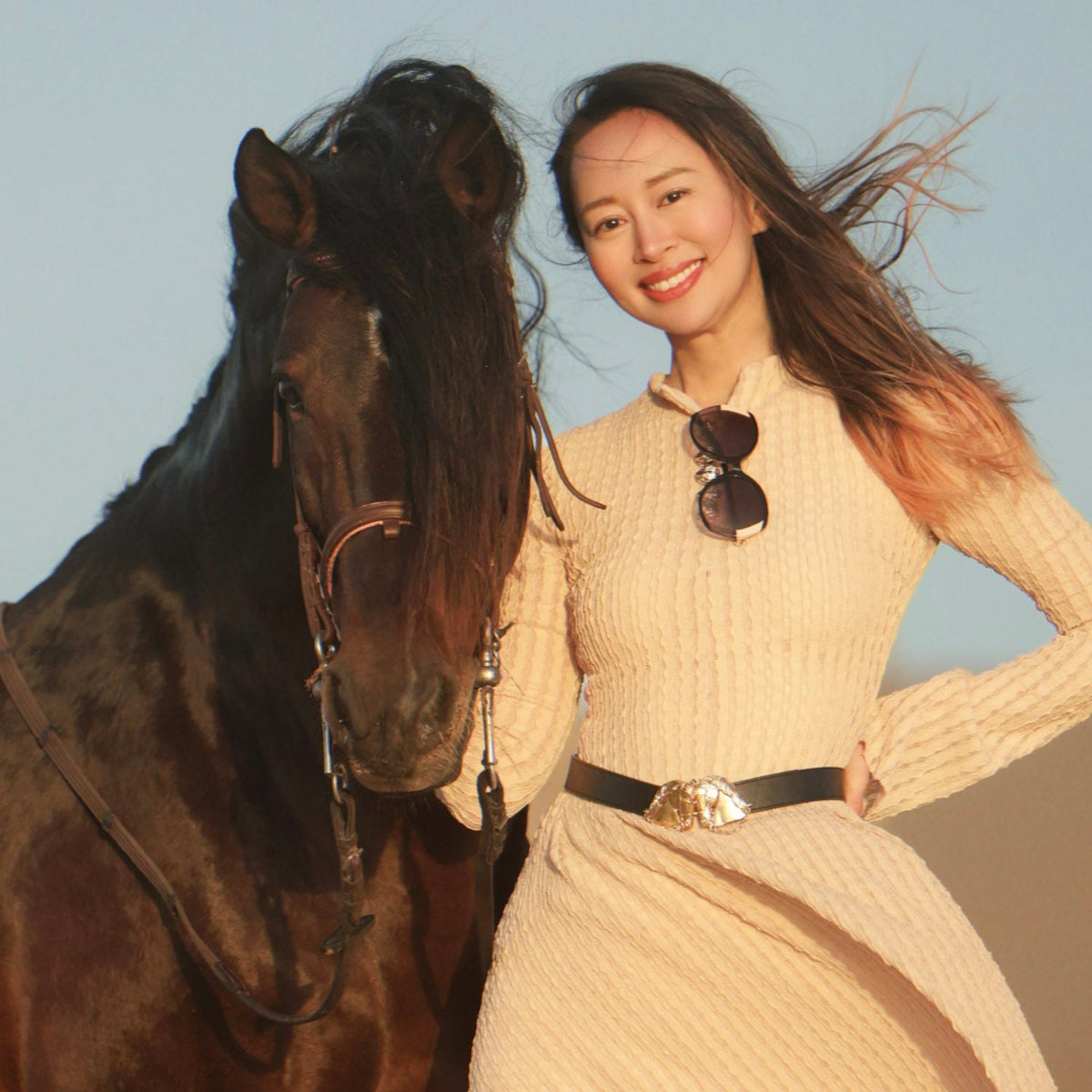 WJY in a beige dress standing next to a horse with a clear sky background